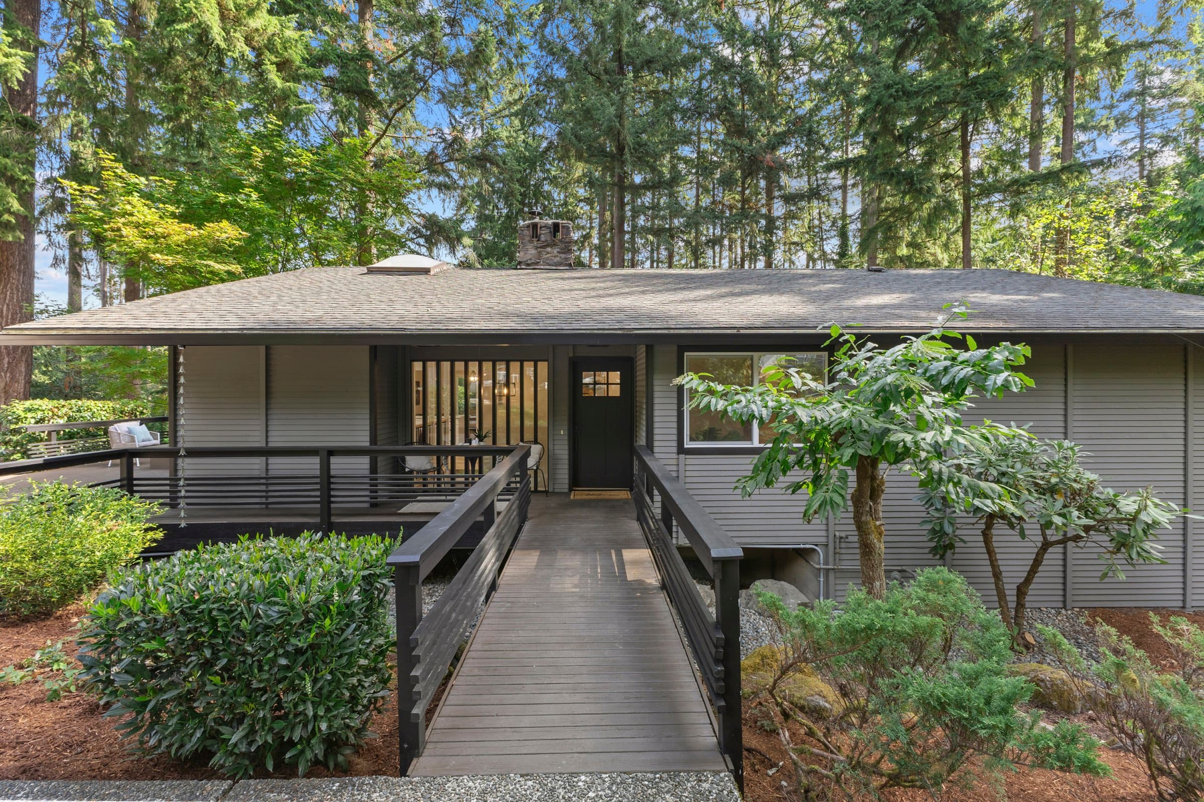 The house from the lawn: a single-storey Northwest home with a board entry deck, under conifers.