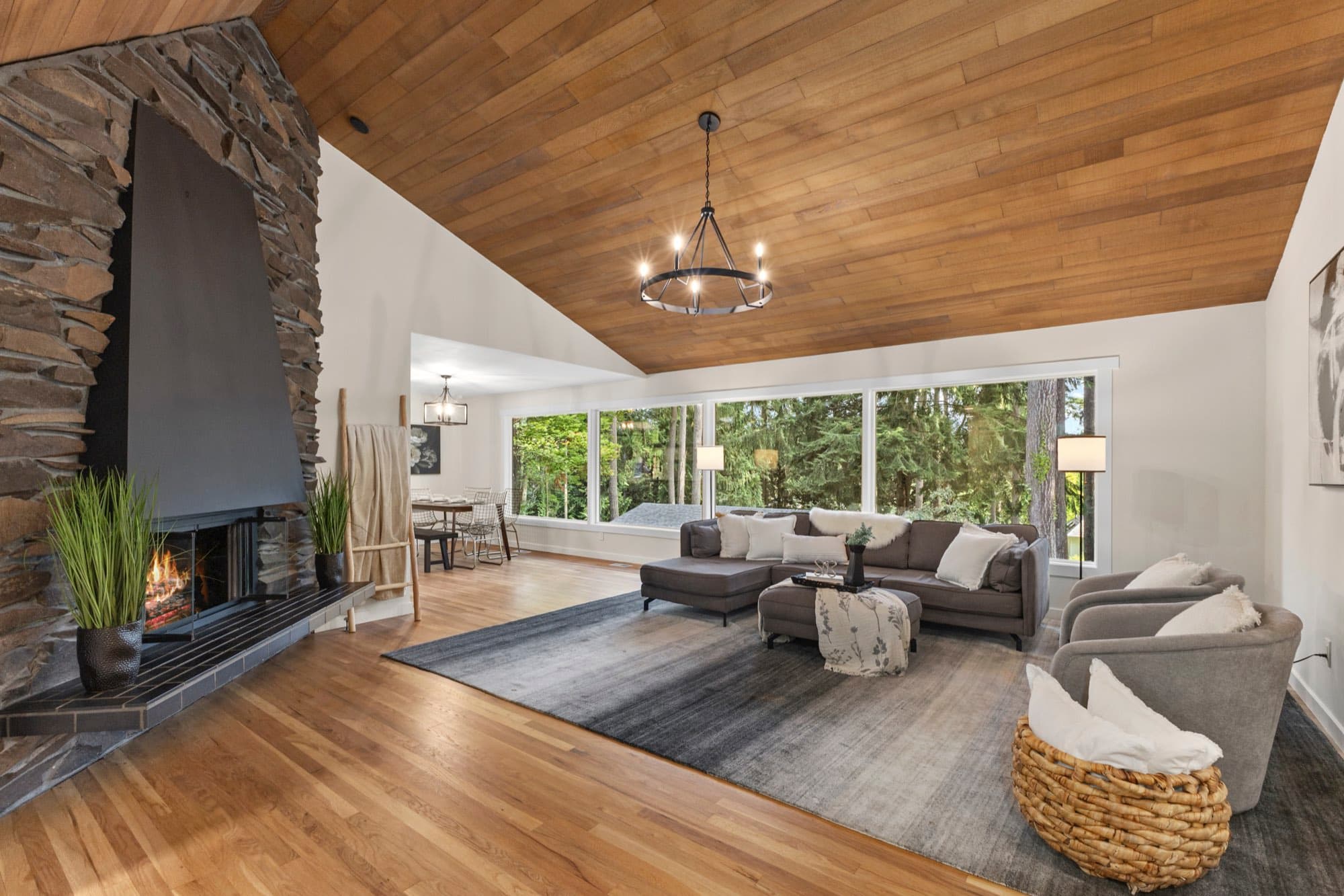 The finished great room: a wood-plank cathedral ceiling, a full-height stone fireplace and a band of windows onto conifers.