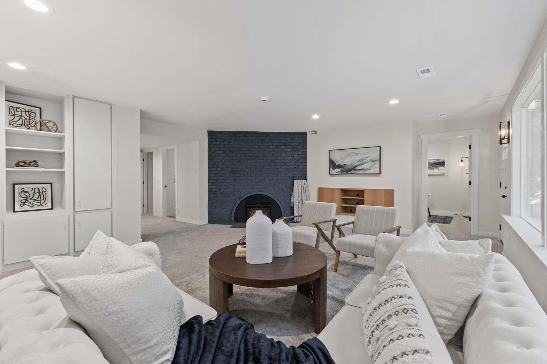 A staged sitting room, pale and bright, with a dark fireplace and built-in shelving.