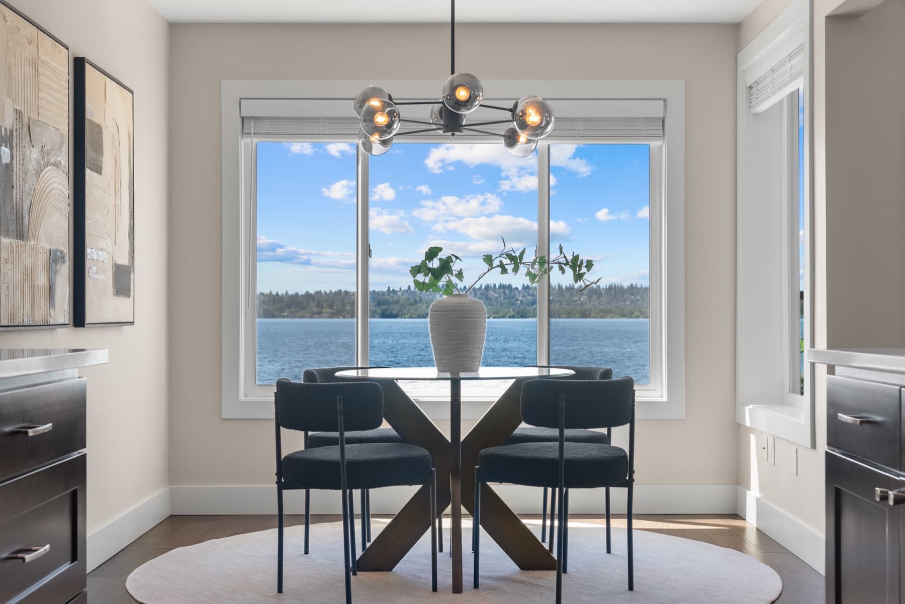 A round dining table and four chairs under a globe pendant, framed by a window onto open water.