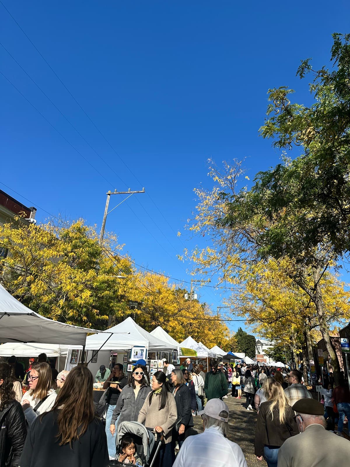 A crowd browsing white market tents lining Ballard Avenue under yellow fall foliage on a Sunday market day.
