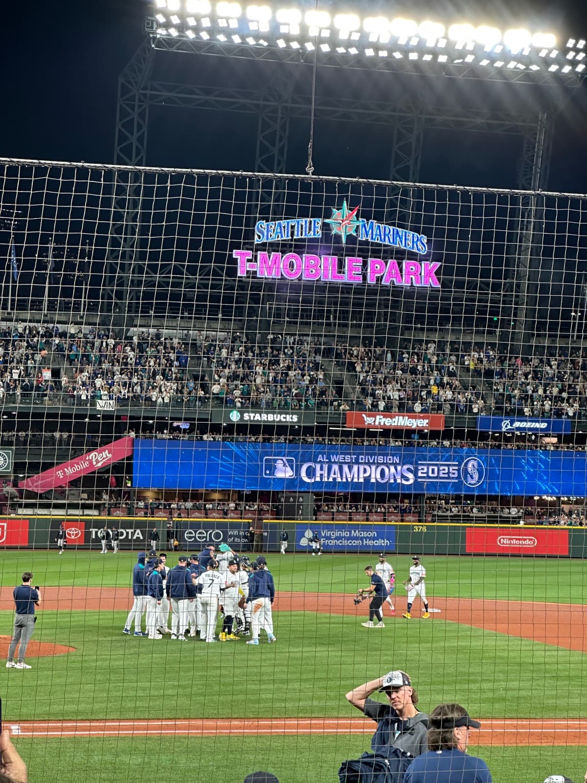 The Mariners celebrating on the field at T-Mobile Park beneath the team's 2025 AL West Champions banner.