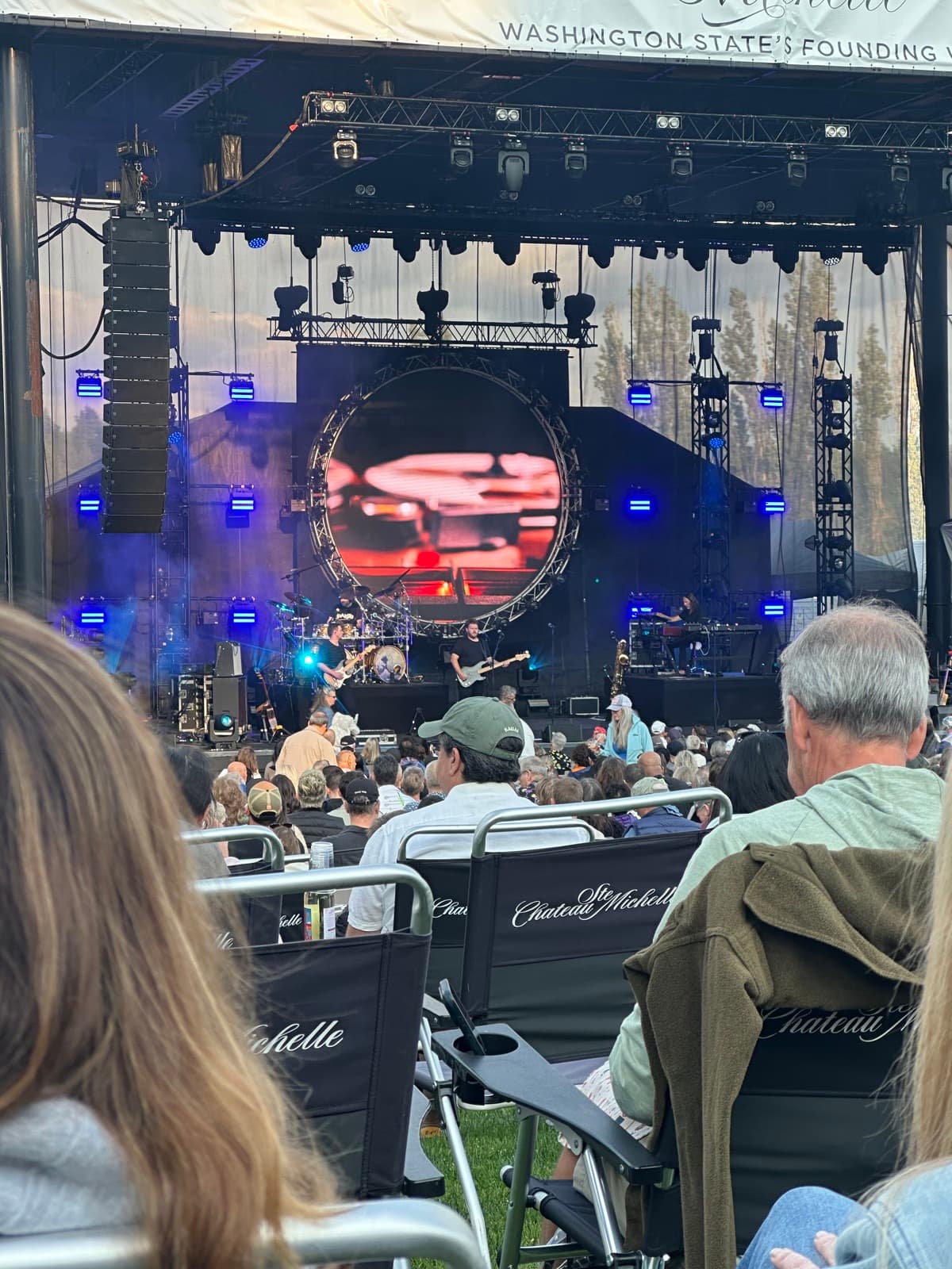 Concertgoers seated in lawn chairs printed with the Chateau Ste. Michelle name, facing a lit outdoor stage with a large video screen at a summer evening show in Woodinville.